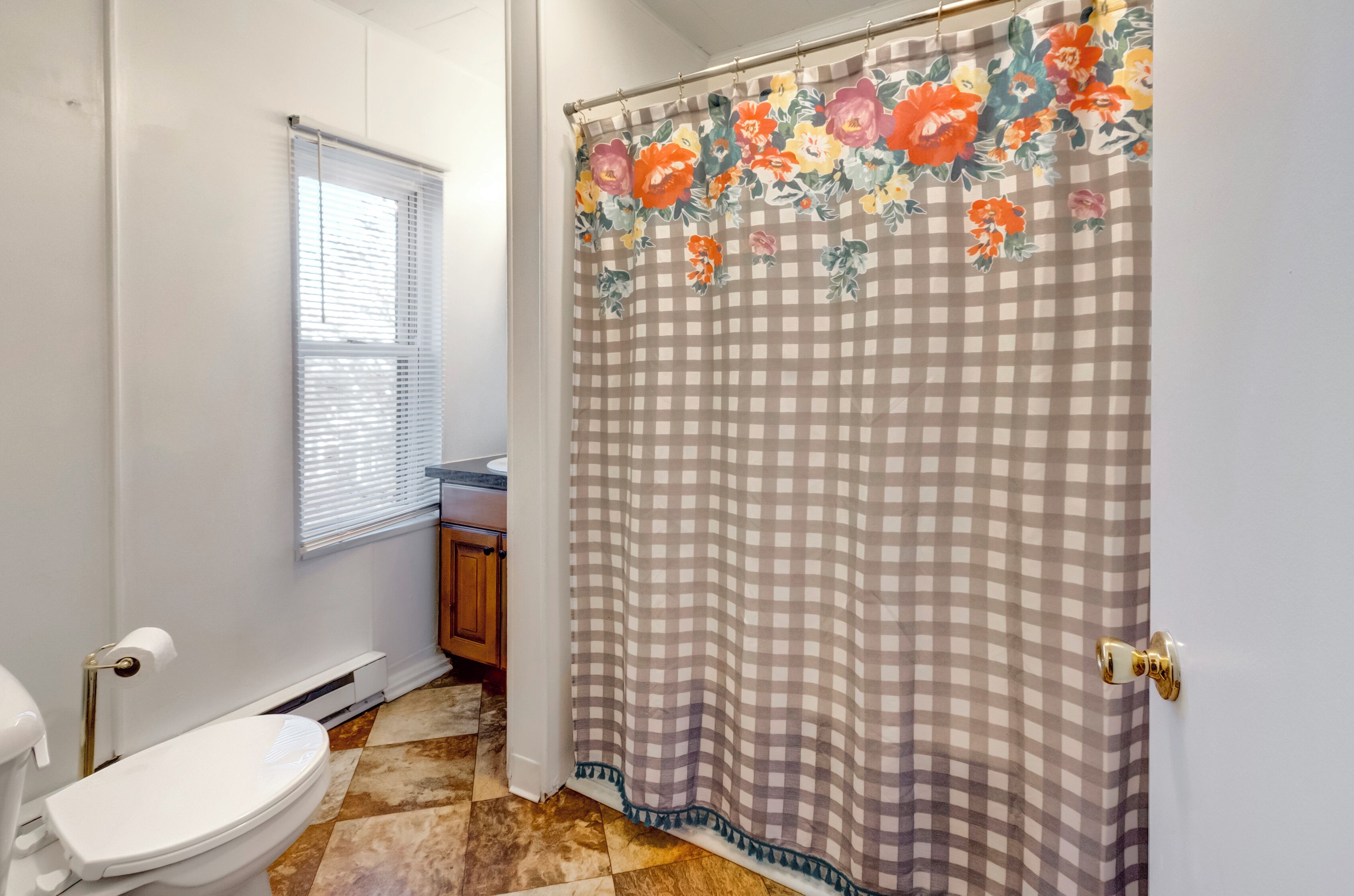 Une salle de bain avec des toilettes, une fenêtre avec des stores, un rideau de douche à motifs floraux et un sol carrelé.