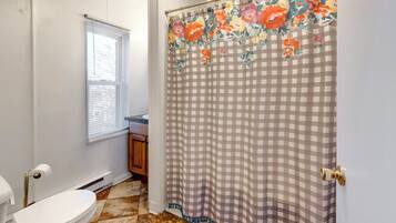 Une salle de bain avec des toilettes, une fenêtre avec des stores, un rideau de douche à motifs floraux et un sol carrelé.