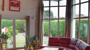 Un salon spacieux avec un canapé en cuir rouge, un plafond en bois, de grandes fenêtres et une vue sur le jardin.