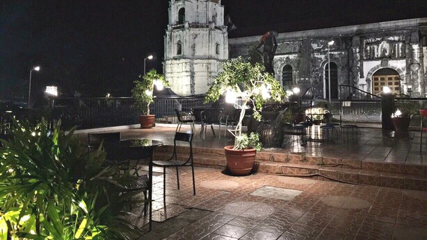 A plaza with a historic building featuring a bell tower, outdoor seating, and potted plants.