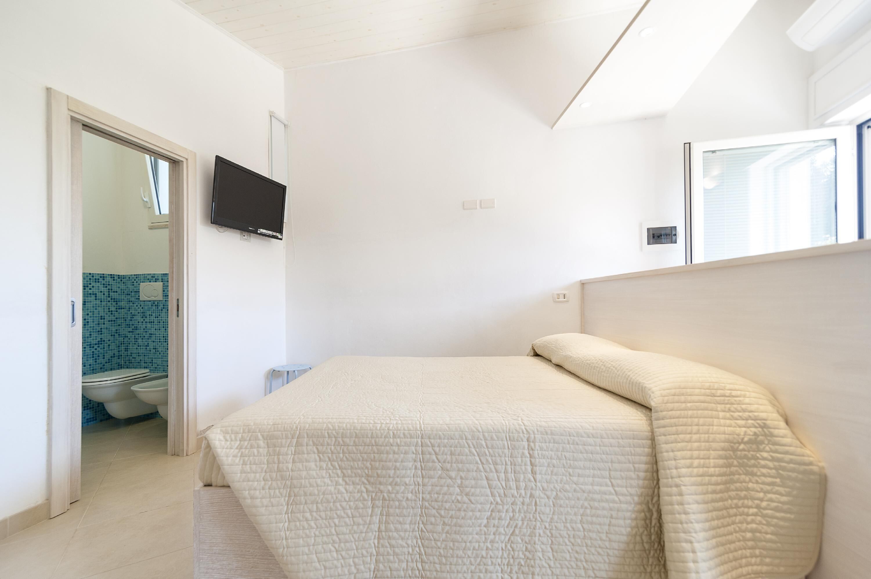 A bedroom with a bed, a TV mounted on the wall, and a bathroom visible through an open door.