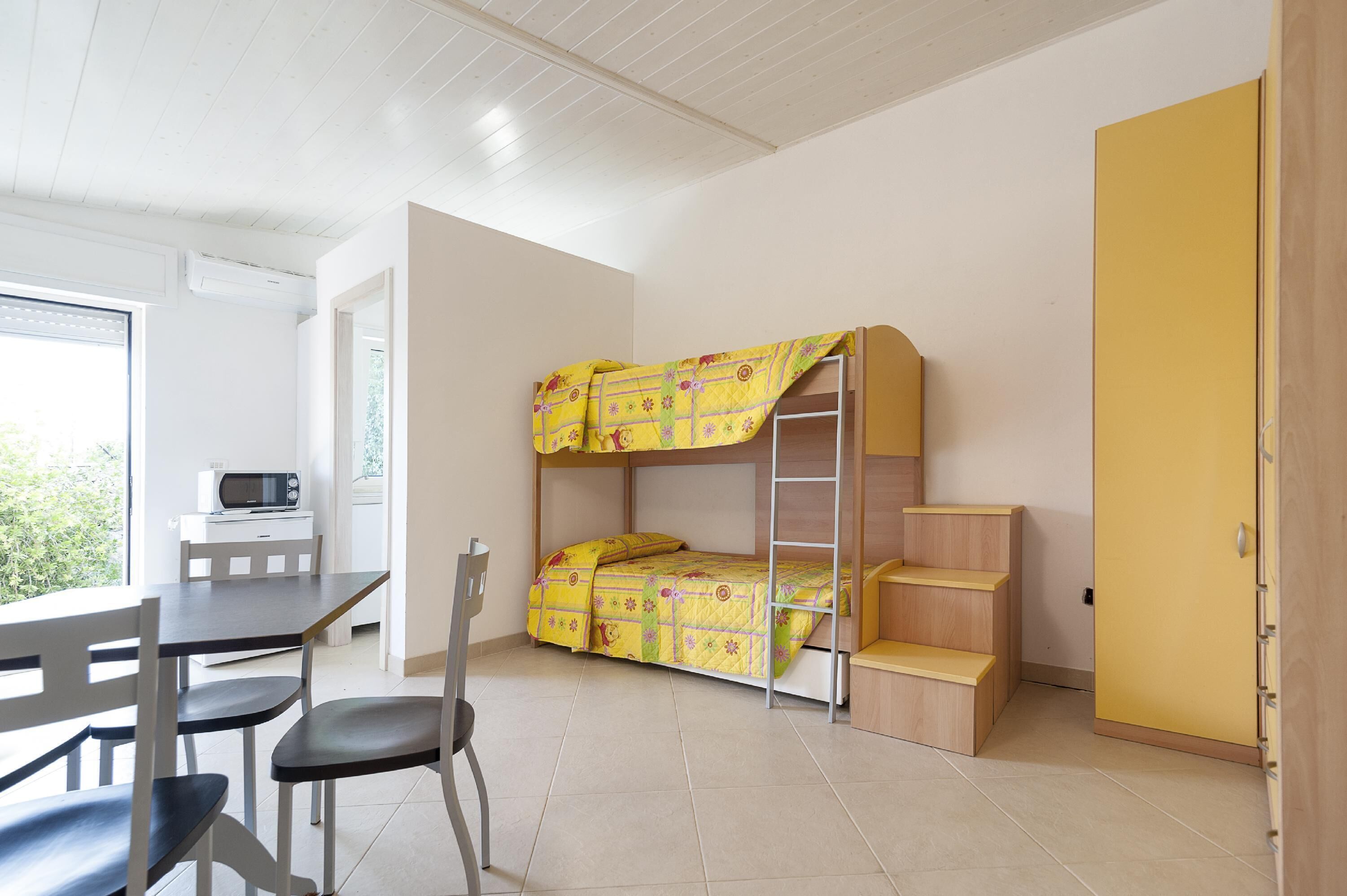 Un lit superposé avec une échelle, un bureau avec une chaise, un four à micro-ondes et une armoire jaune dans une pièce aux murs blancs et au sol carrelé.