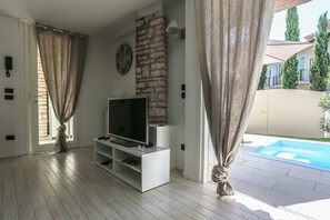 Ein Wohnzimmer mit Fernseher auf einem weißen Schrank, einer Steinwand und Blick auf einen Swimmingpool.