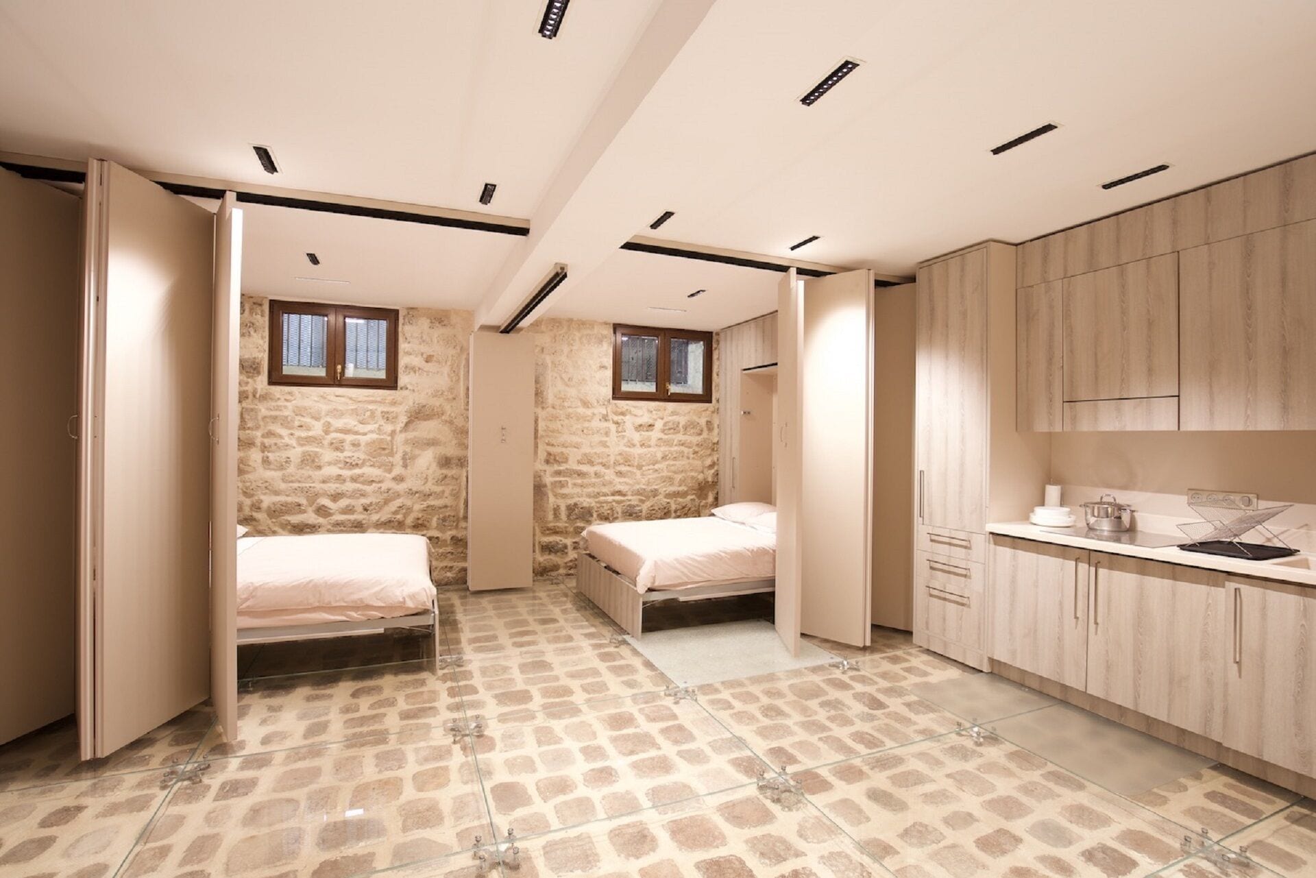 A compact room with two beds, a kitchenette, and a stone wall.