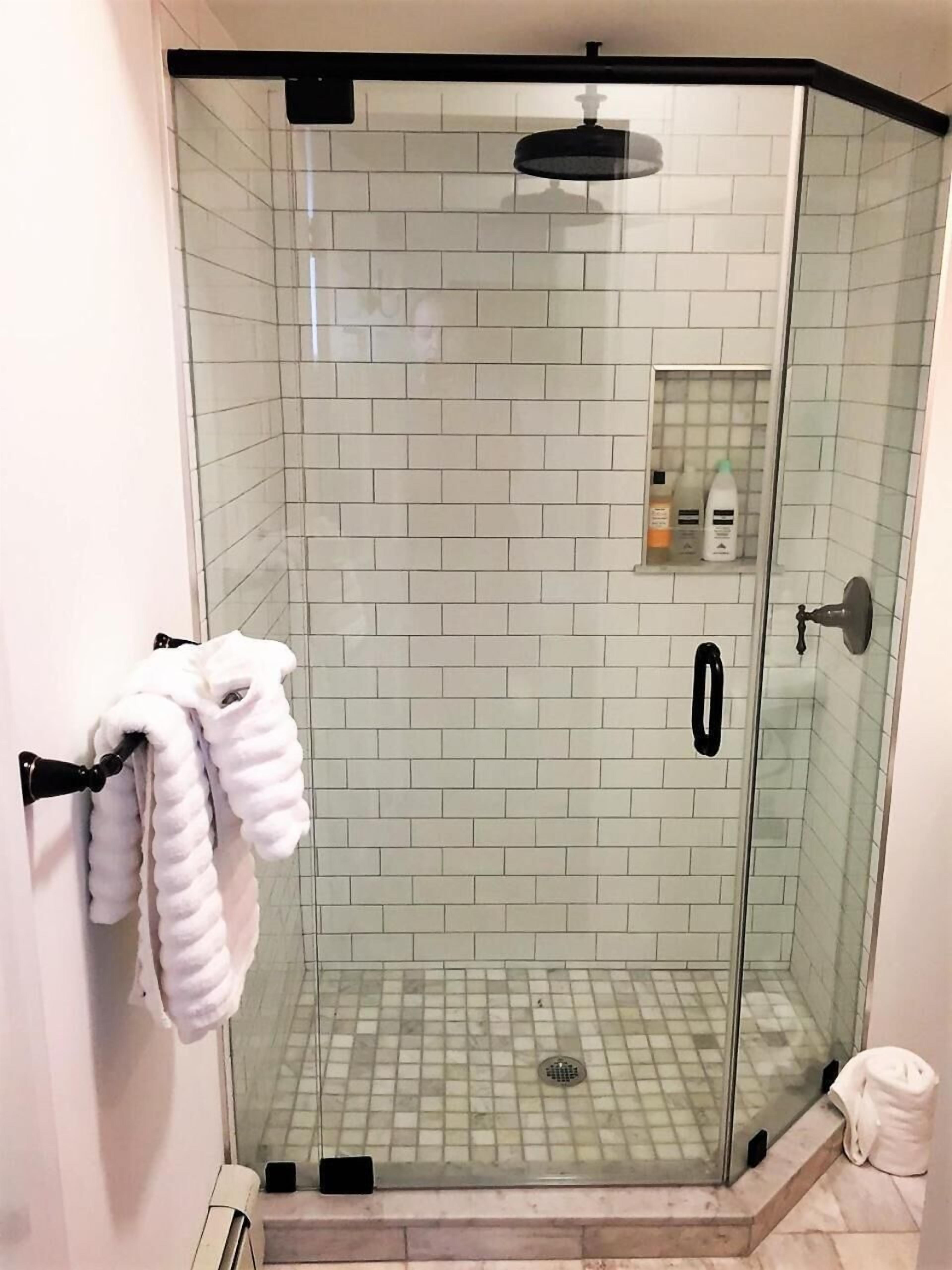 A glass-enclosed shower with white tiles, a towel rack with towels, and a small shelf with toiletries.