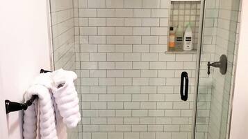 A glass-enclosed shower with white tiles, a towel rack with towels, and a small shelf with toiletries.