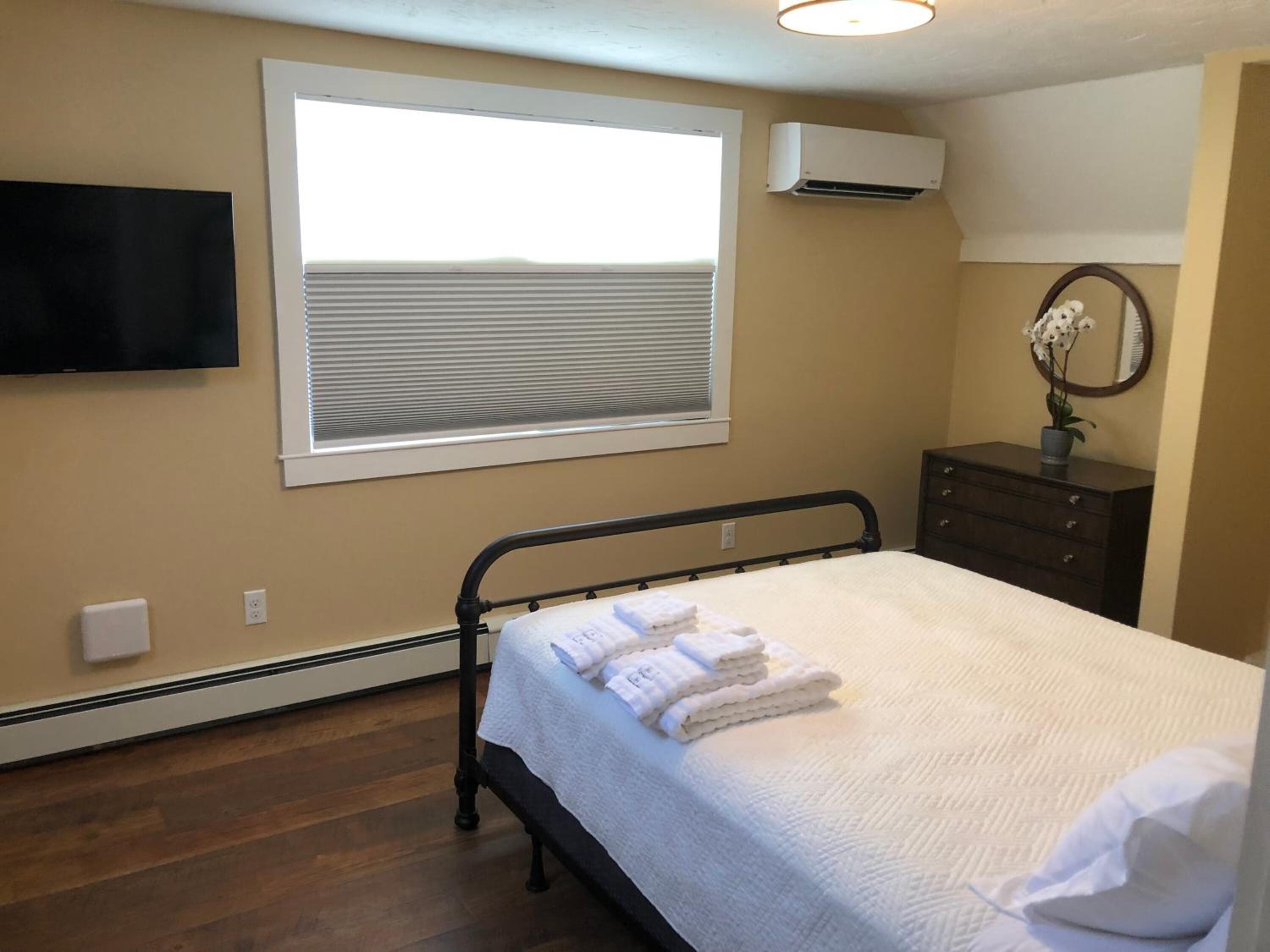 Une chambre à coucher comprenant un lit, une fenêtre avec des stores, un climatiseur, une commode et un téléviseur à écran plat fixé au mur.