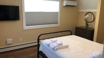 Une chambre à coucher comprenant un lit, une fenêtre avec des stores, un climatiseur, une commode et un téléviseur à écran plat fixé au mur.