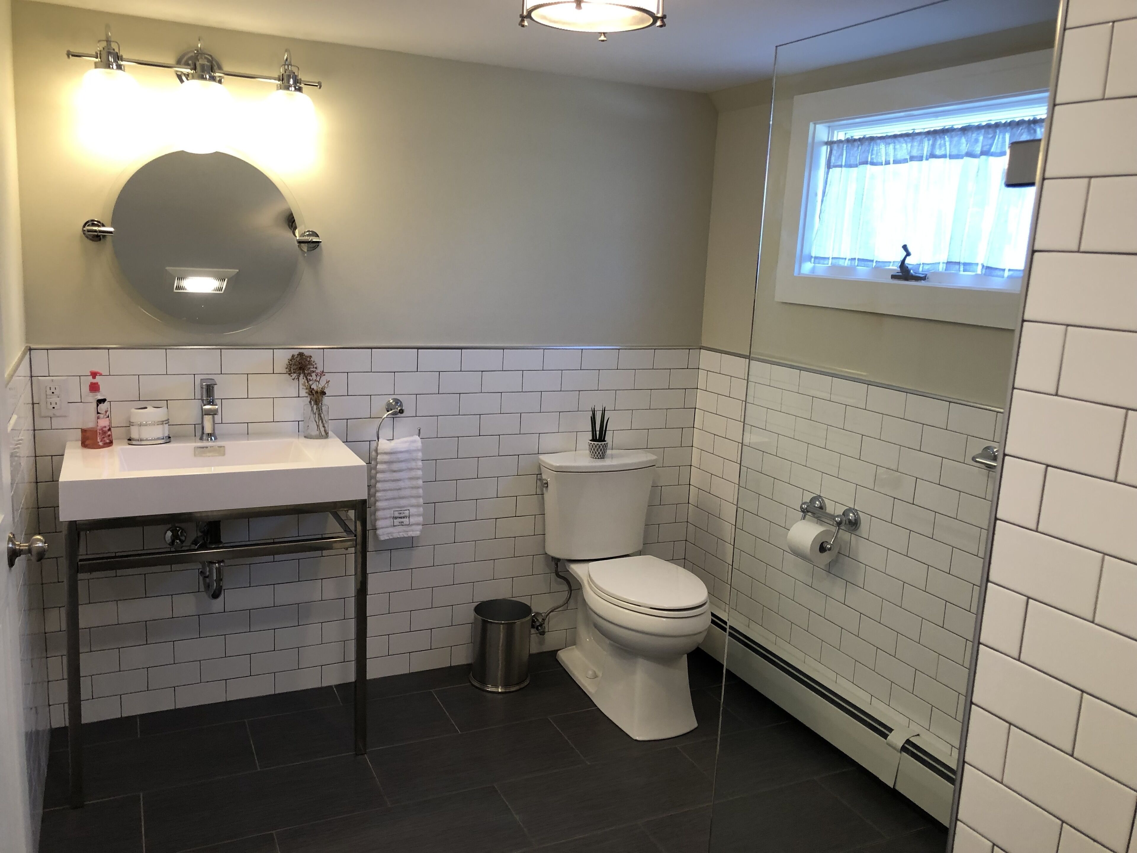 Ein modernes Badezimmer mit Waschbecken, Spiegel und Toilette in Weiß. Ein Mülleimer und ein Fenster mit Vorhängen sind ebenfalls vorhanden.