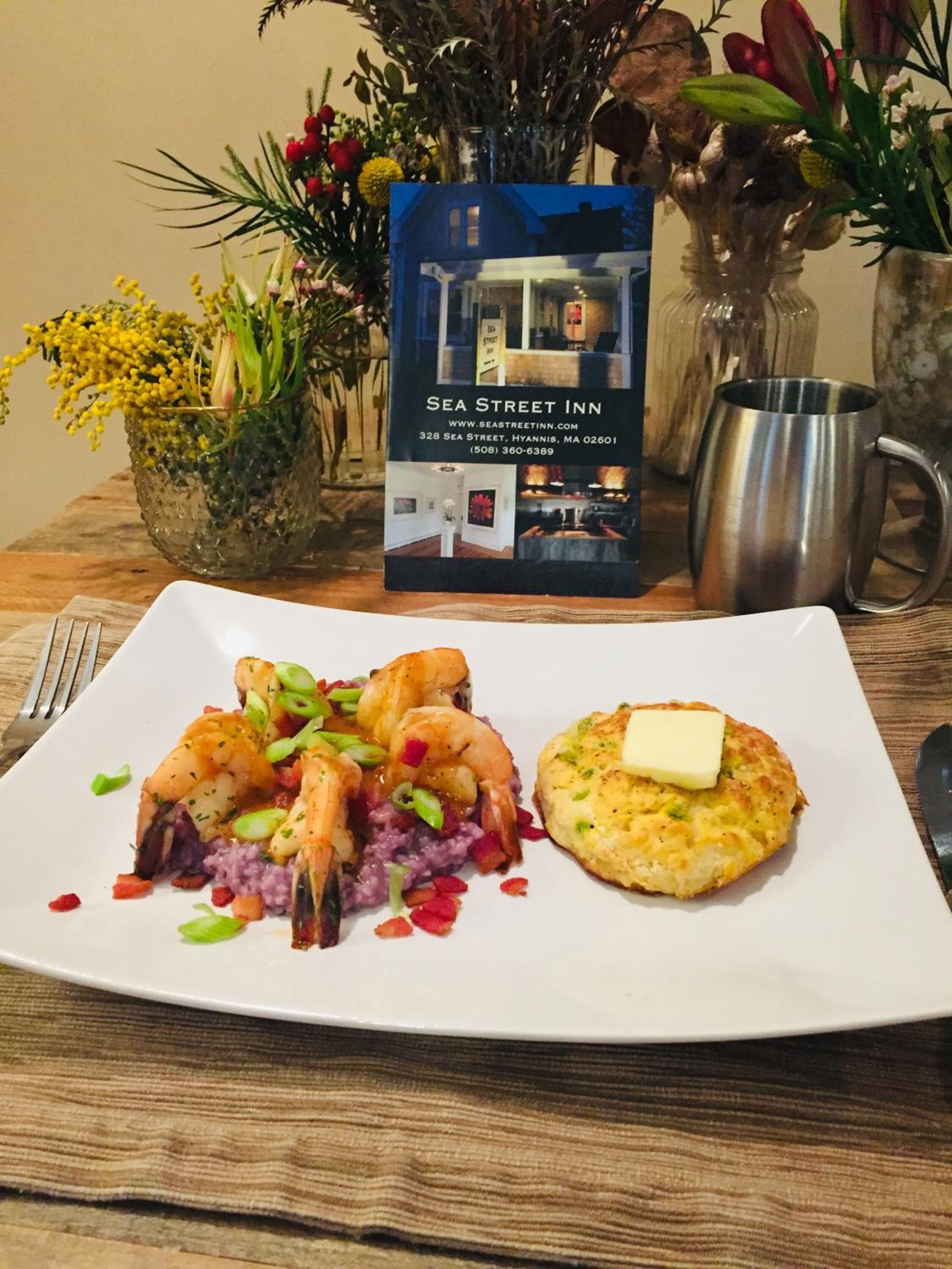 En tallerken med rejer og grits med en smørstegt kiks på et træbord. En brochure for Sea Street Inn er synlig i baggrunden.