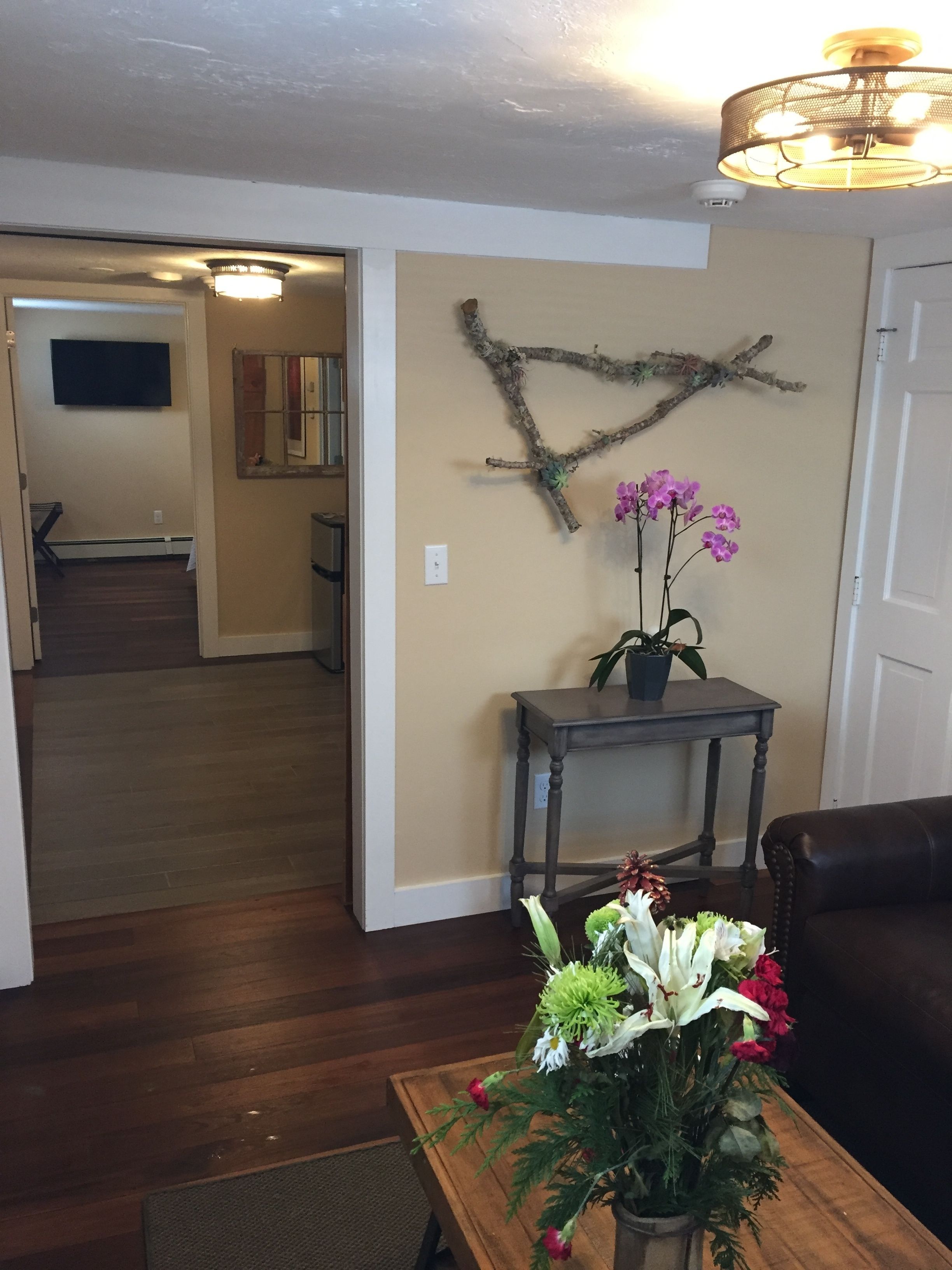 A living room with a wooden floor, a dark brown leather sofa, a coffee table with a floral arrangement, a wall-mounted branch decoration, and a ceiling light fixture.