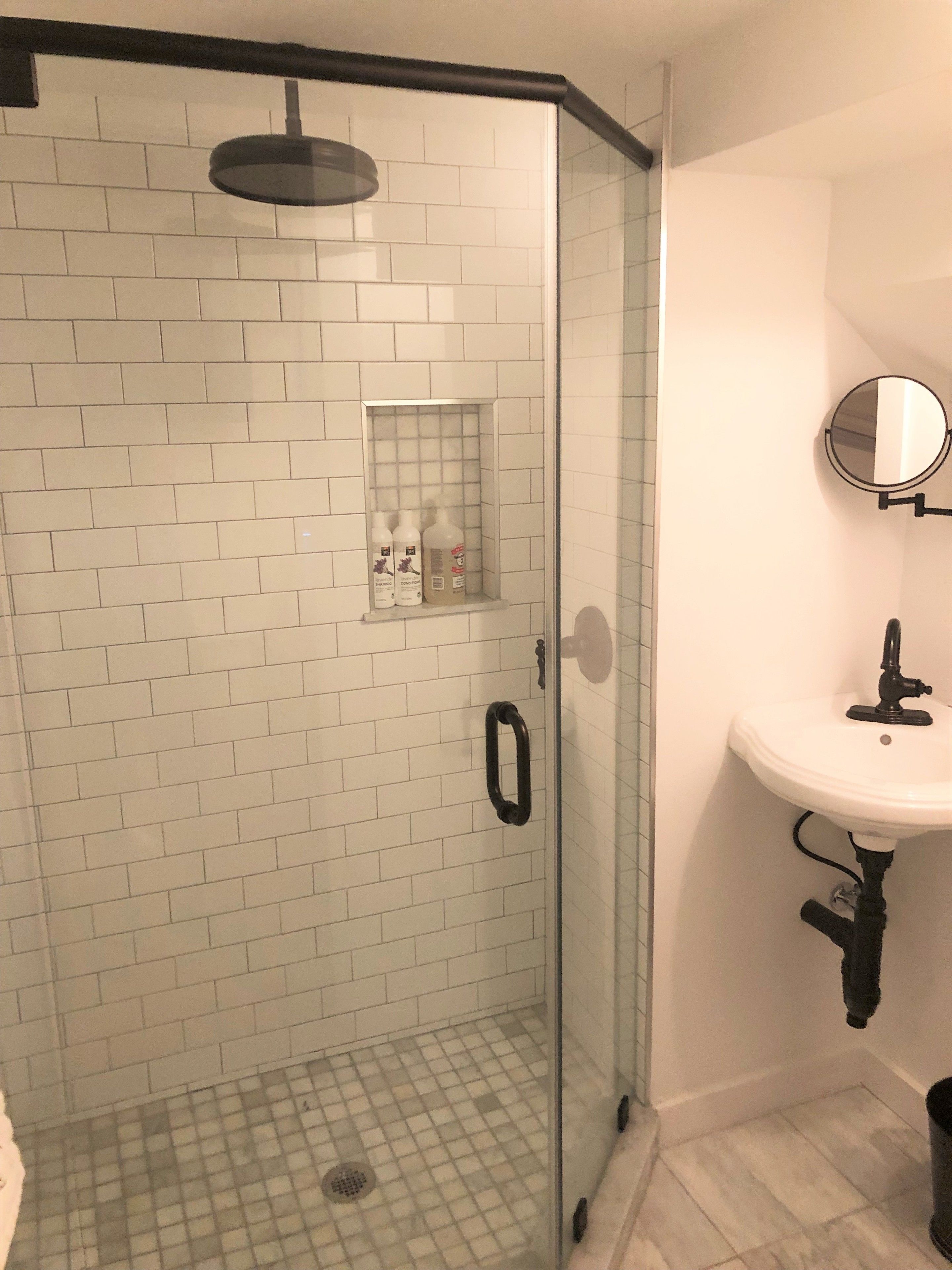 Une douche fermée par des parois de verre, avec des carreaux de céramique blancs rectangulaires, une porte en verre et une petite étagère pour les articles de toilette.