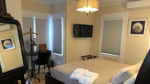 A neatly arranged bedroom with a large bed, a bedside table, a chair, a TV mounted on the wall, and a window with blinds. - Sea Street Inn (Hyannis)