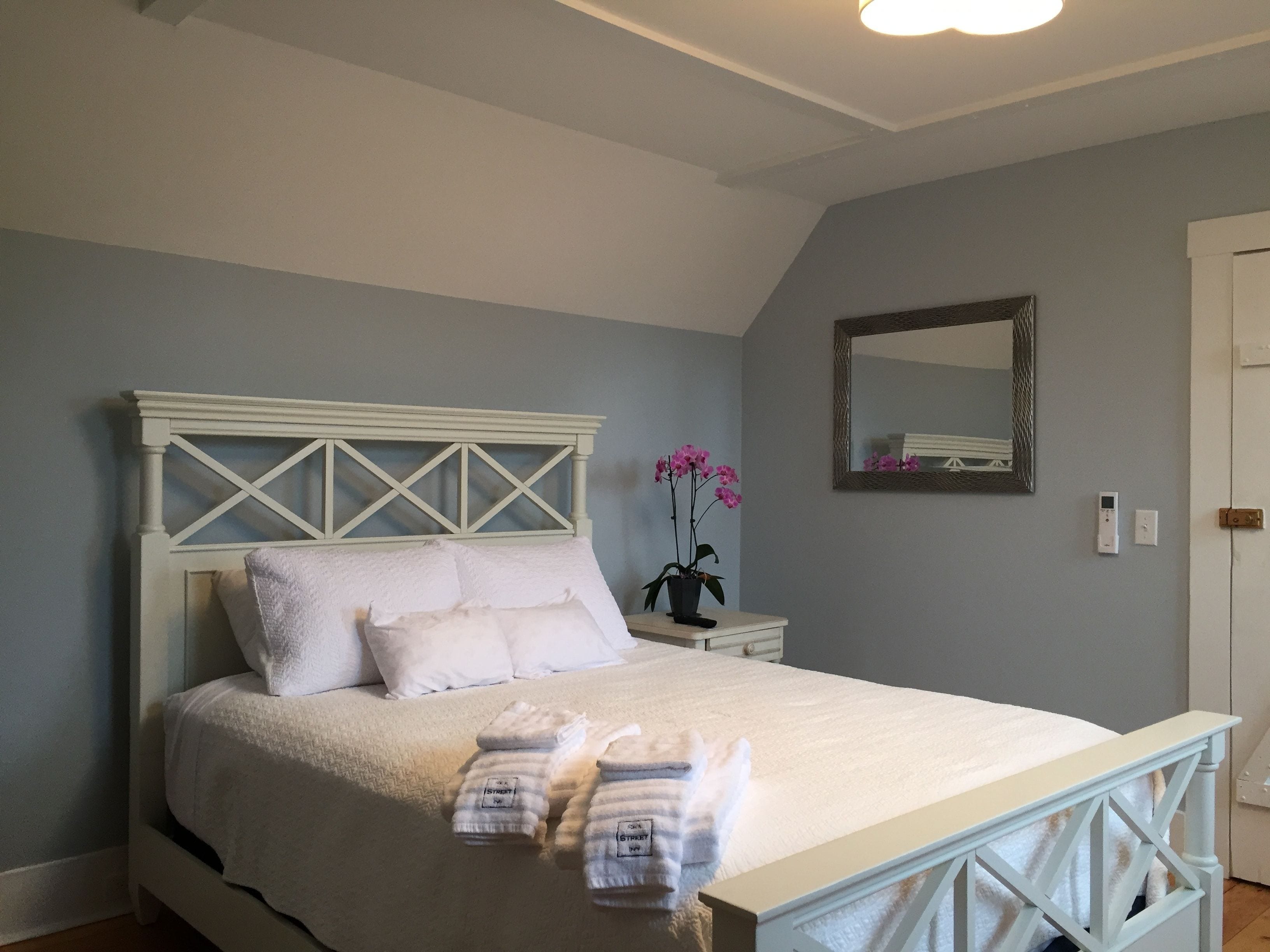 Un lit bien fait, avec du linge de lit blanc et une tête de lit, une orchidée en pot et une table de chevet avec un petit miroir et un thermostat.