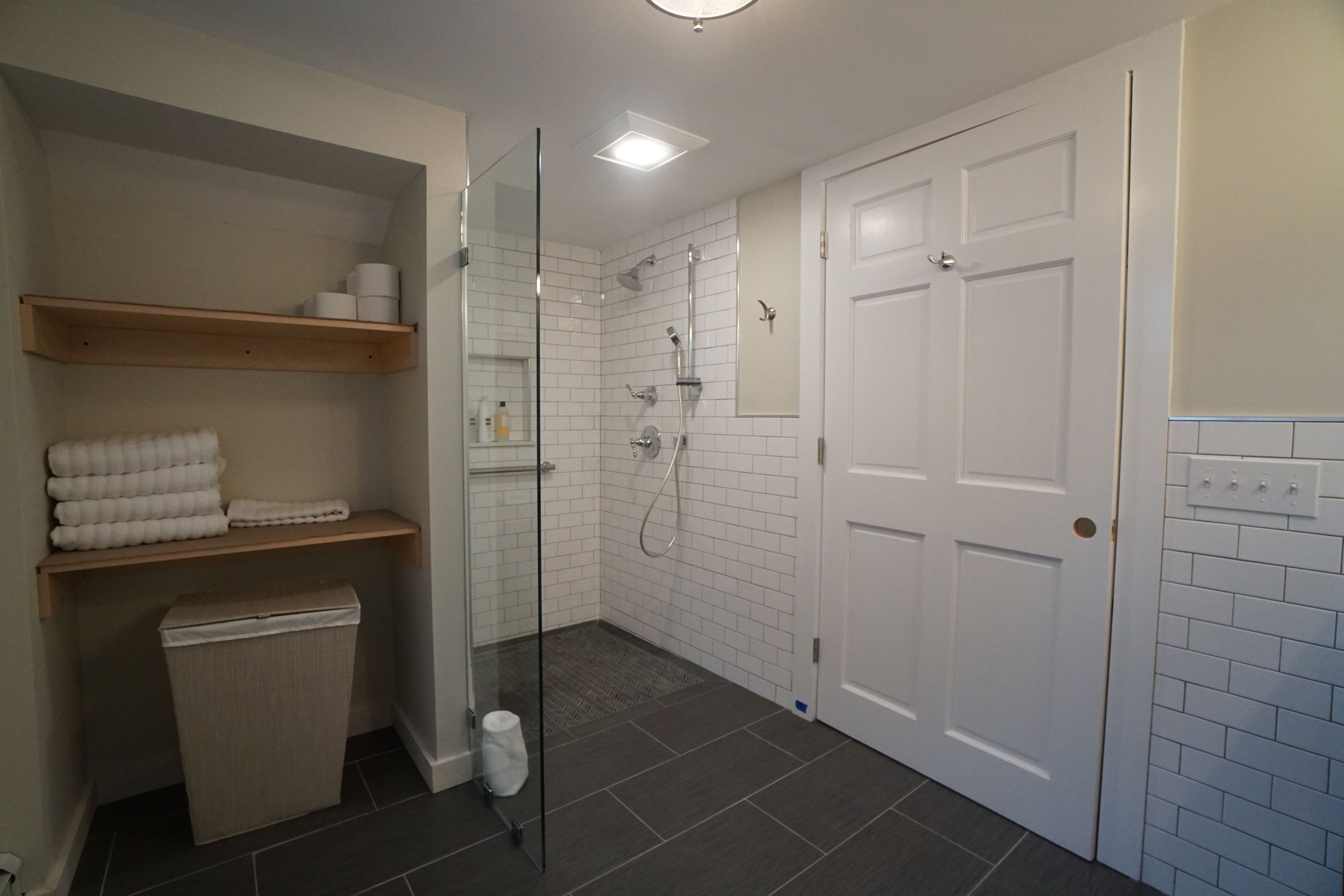 A modern bathroom with a glass-enclosed shower, white tiled walls, and a wooden shelf with towels.