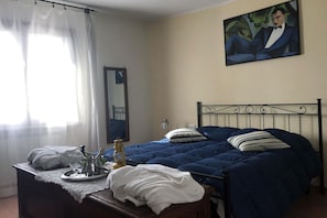 A bedroom with a bed, a bedside table with a tray of items, a window with curtains, and a painting on the wall. - Apartment with pool use, terrace, historical centre, for family (sermoneta)