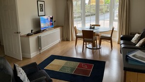 A living room with a television, a dining table with chairs, a sofa, and a view of the outdoors through large windows.