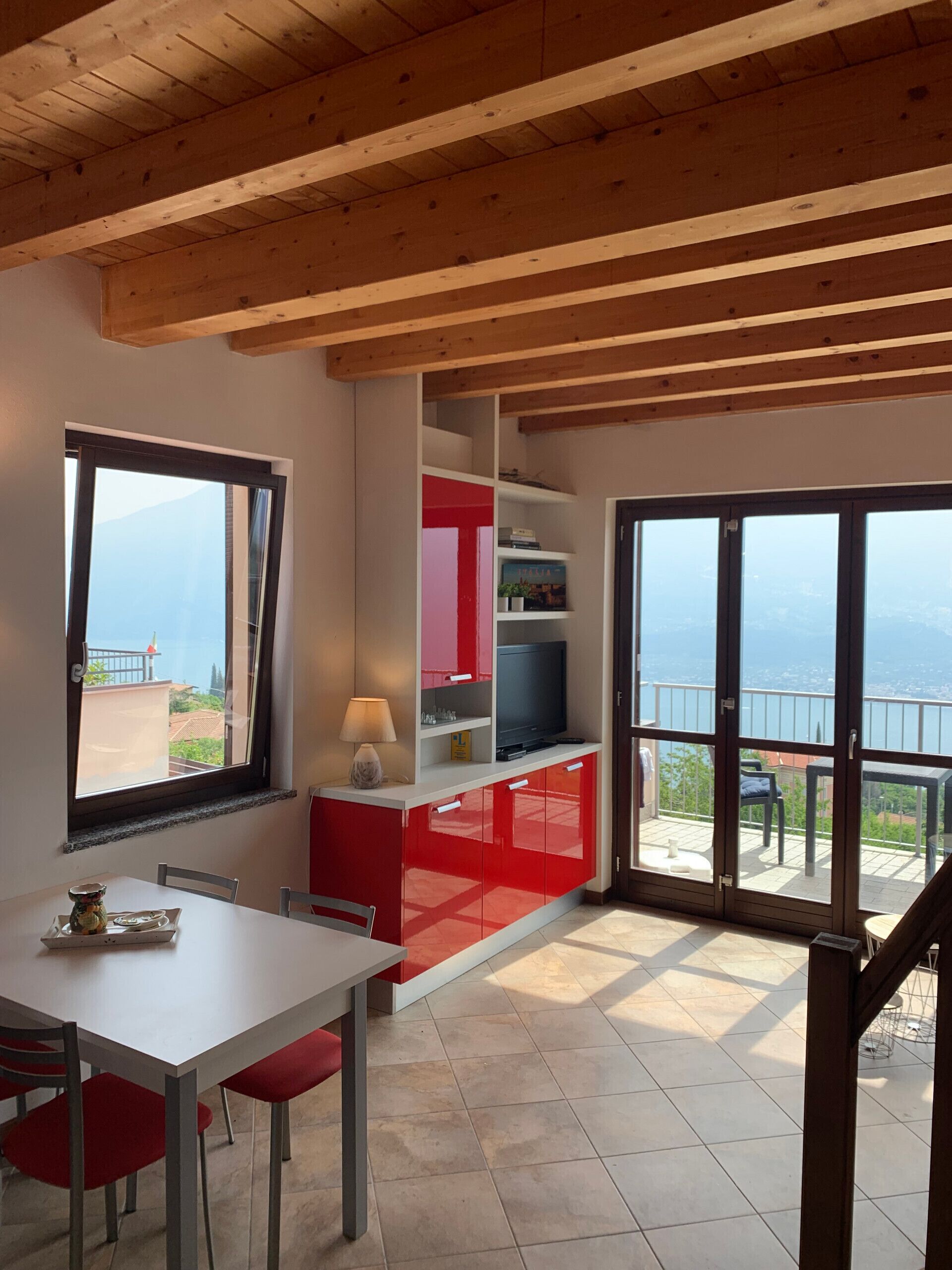 Une pièce comprenant une table à manger et des chaises, un téléviseur posé sur un meuble rouge, et un balcon avec vue.