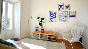 A modern living room with a glass coffee table, a wicker ceiling fan, a white chair, and a bookshelf with framed artwork. - Charming Graça - 4 pax (Lisboa)