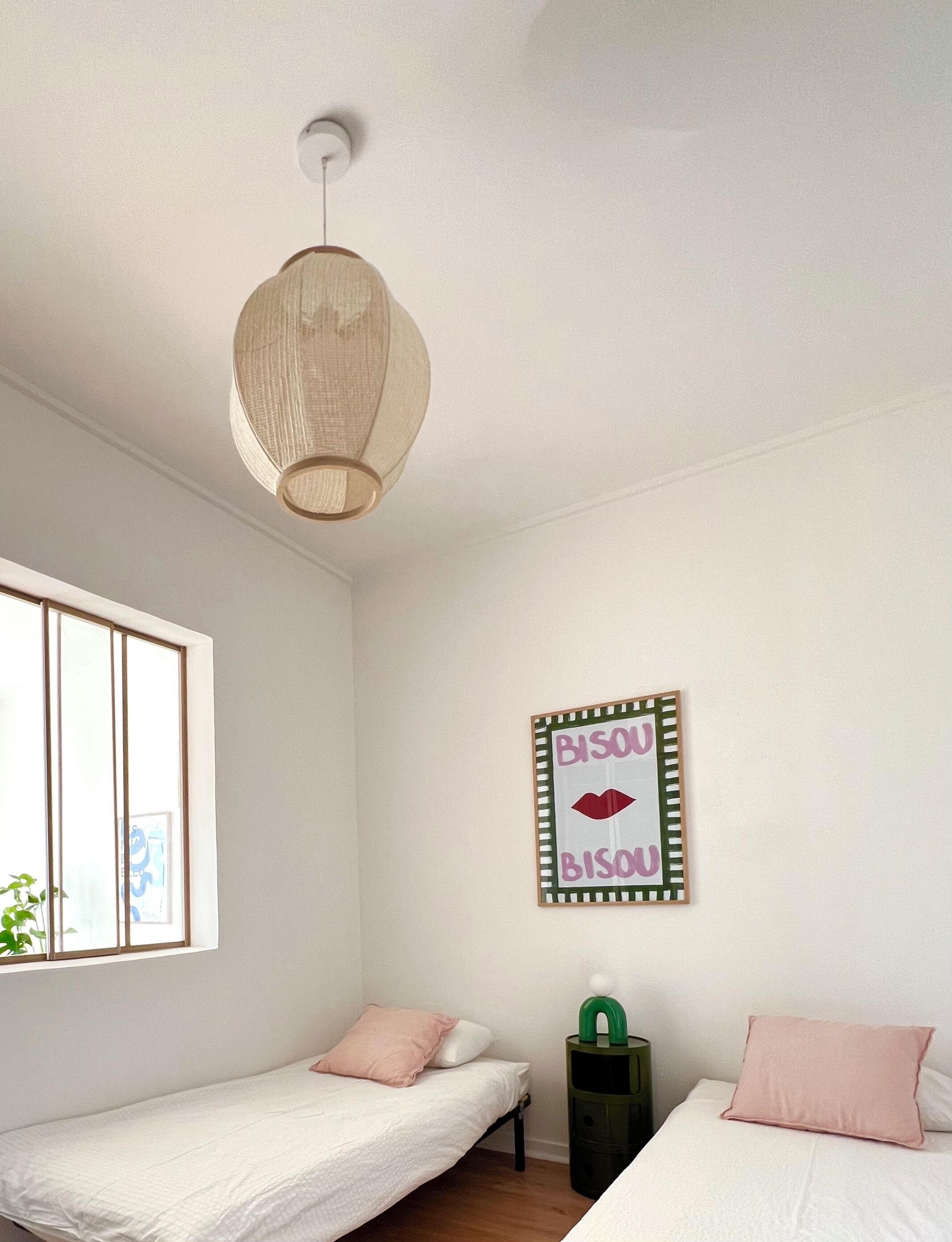 A bedroom with two single beds, a hanging light fixture, a framed sign on the wall, and a window with blinds.