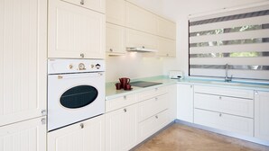 Een keuken met witte kastjes, een inbouwoven, een raam met zonwering en een aanrecht met een waterkoker en koffiezetapparaat.
