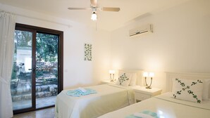 Hotelkamer met twee bedden, een plafondventilator, een airconditioning en uitzicht door een glazen deur op een buitenruimte met een stenen muur en groen.