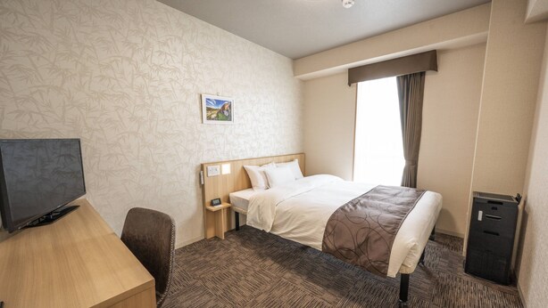 A hotel room with a bed, a desk with a computer, a TV, a small bedside table, and a waste bin. - Tabino Hotel Sado (Sado)