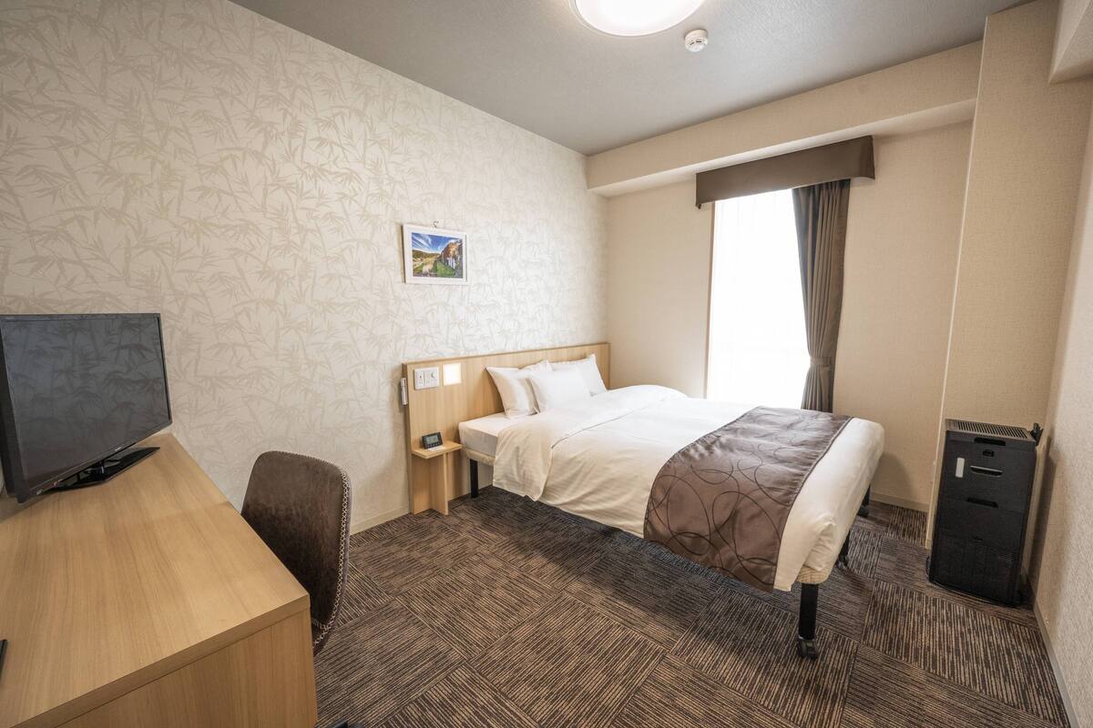 A hotel room with a bed, a desk with a computer, a TV, a small bedside table, and a waste bin.