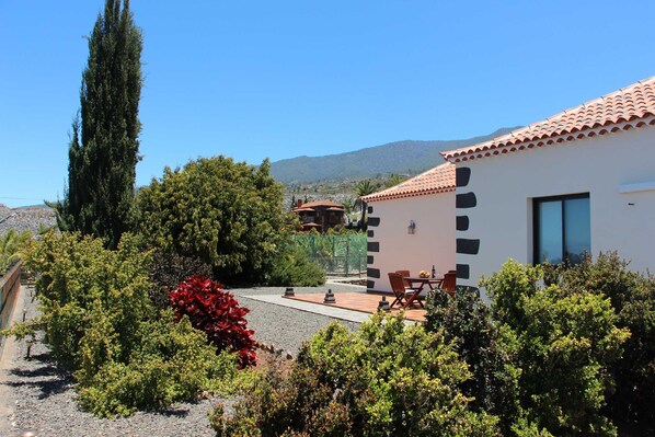 A modern building with a tiled roof, a patio with outdoor furniture, and a garden with various plants and a tall tree. - Casa Luque, La Punta, Spain (Tijarafe)