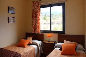 A bedroom with two beds, a window with a view, a nightstand with a lamp, and two framed pictures on the wall.