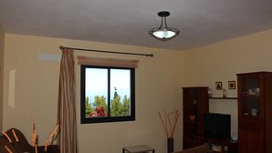 A living room with a television, a small table, a sofa, a window with a view of trees, and a ceiling light. - Casa Luque, La Punta, Spain (Tijarafe)