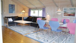 Una habitación acogedora con un sofá de cuero negro, una mesita pequeña y sillas coloridas. Se ve una ventana con cortinas moradas y un cuadro en la pared.
