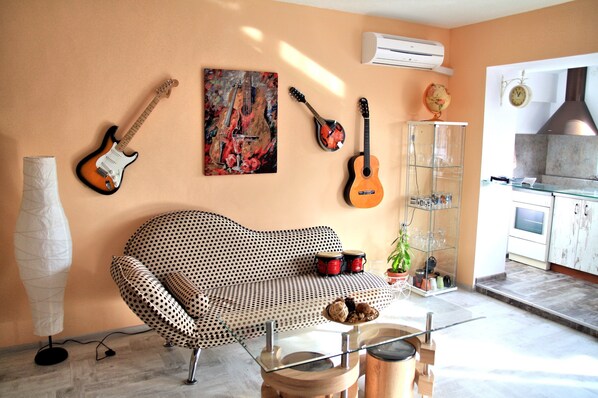 A living room with a patterned sofa, a glass coffee table, a guitar, a ukulele, a globe, and a kitchen area with cabinets and a stove.