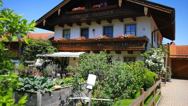 A traditional chalet with a wooden balcony, a garden with various plants, and a clear blue sky. - Ferienwohnung II° "Bergstüberl" mit extra Schlafzimmer und Balkon, 60 qm (Aschau im Chiemgau)