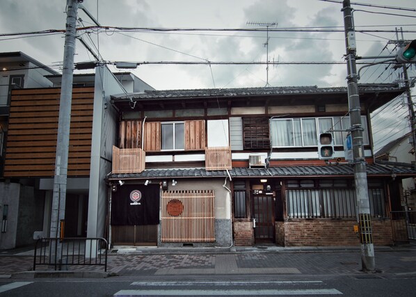 Un bâtiment traditionnel japonais, avec une façade en bois, un toit recouvert de tuiles et une porte noire ornée d’un emblème circulaire.