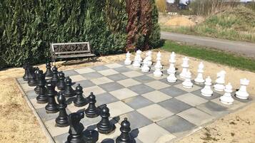 A large outdoor chessboard set up on a paved area with a bench and greenery in the background.