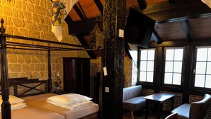 A room with a stone wall, a wooden bed with a headboard, a bench, a table, and a television mounted on the wall.