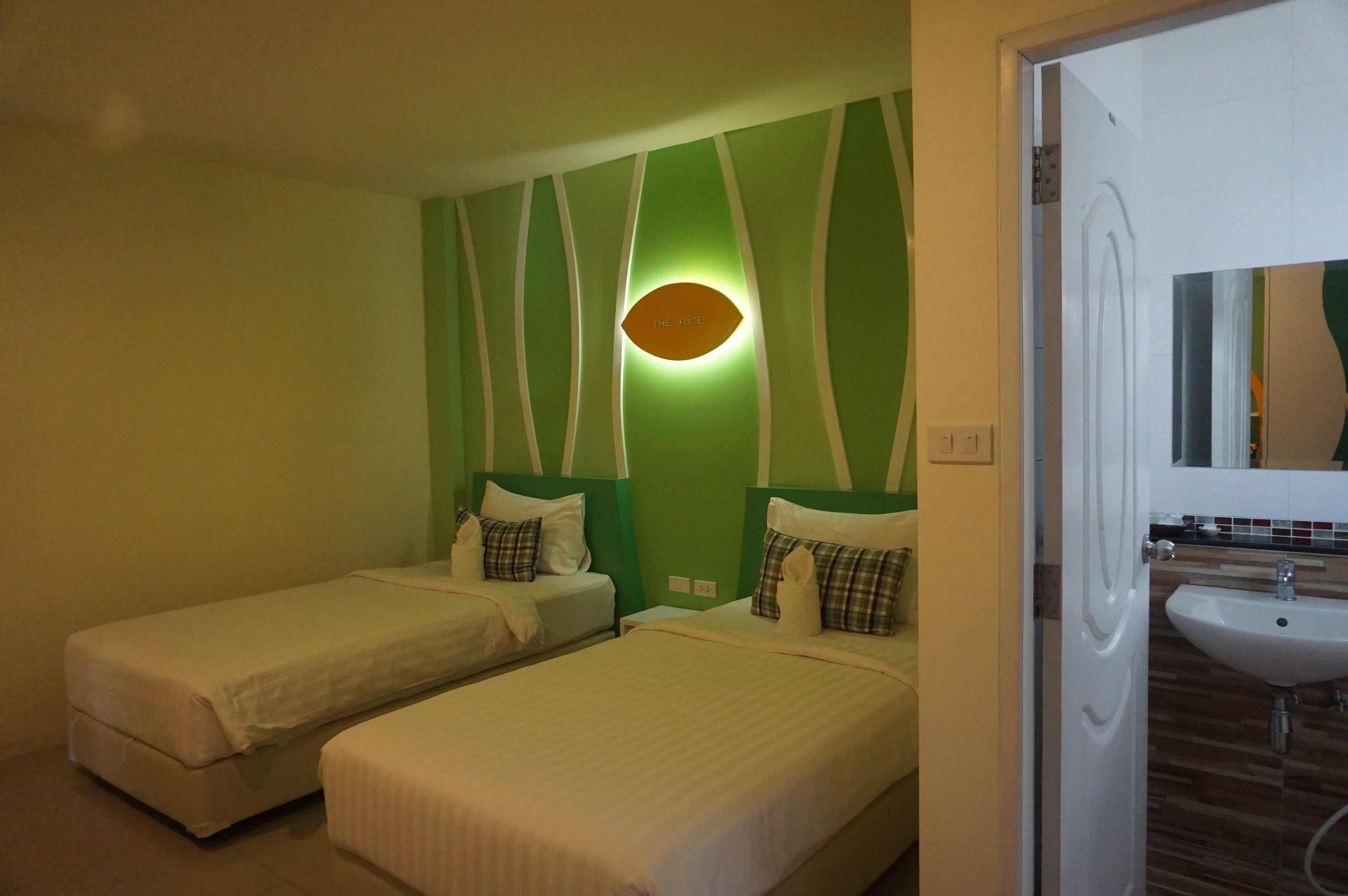A hotel room with two single beds, a green accent wall, a wall-mounted light, and a bathroom with a sink and mirror.