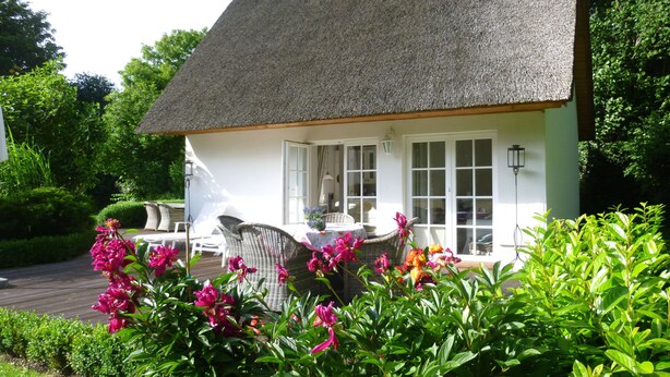 Ein weißes Cottage mit Strohdach, einem Essbereich mit Korbmöbeln und einem Garten mit rosa Blumen.