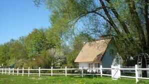 Ein traditionelles Haus mit einem Reetdach, umgeben von einem weißen Zaun, und eine Wassermühle im Hintergrund.
