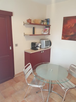 Una cocina compacta con una mesa redonda de vidrio, dos sillas de metal, un microondas, un tostador y estantes con utensilios de cocina.