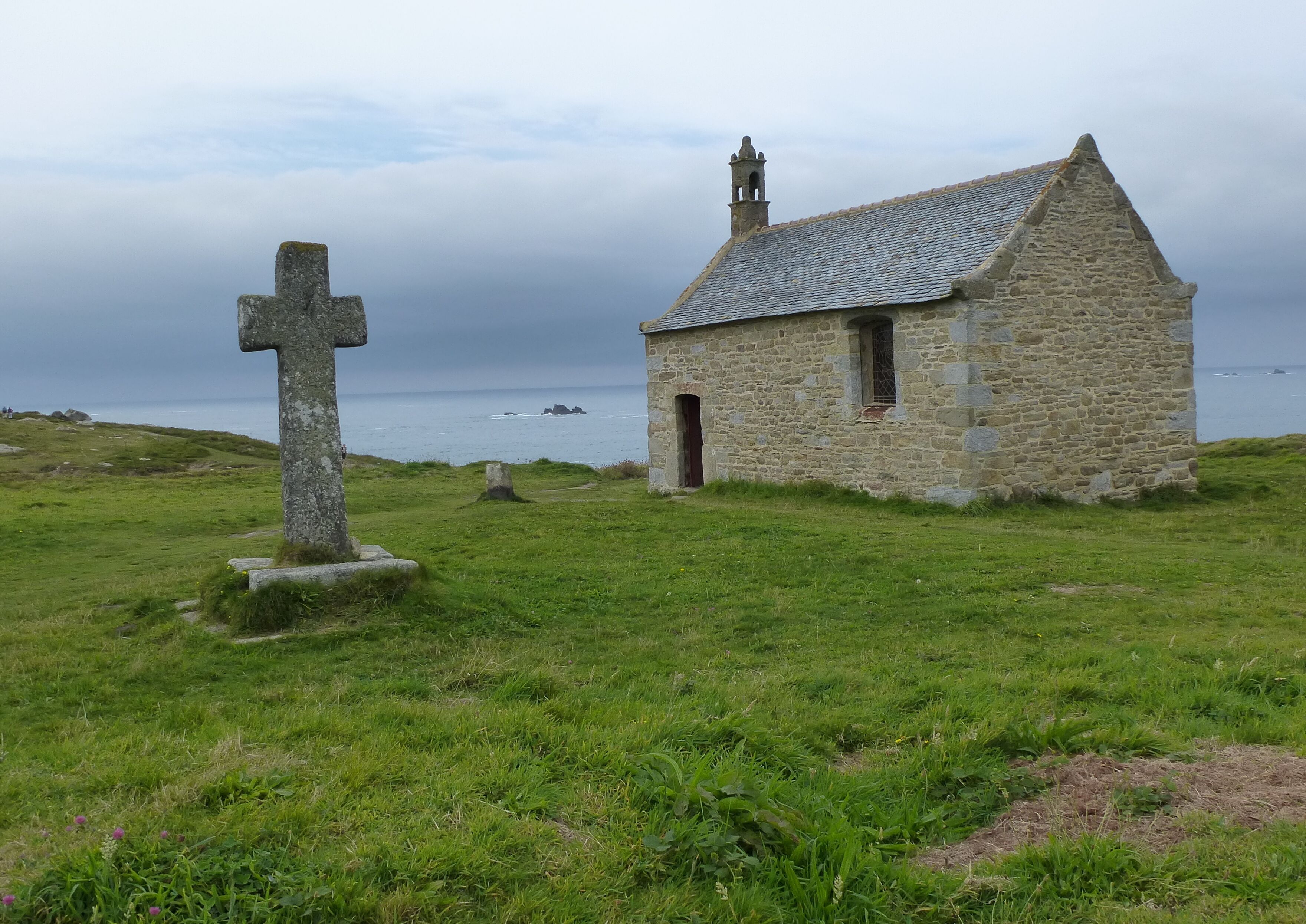 Une église en pierre, dotée d’un clocher et d’une croix, se dresse dans un champ verdoyant.