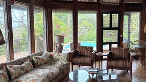 Ein geräumiges Zimmer mit einer großen Couch, zwei Sesseln, einem Glastisch und Blick auf einen Pool durch große Fenster.
