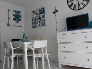 A white dining table with chairs, a white dresser, a clock, and wall decorations including a sign that says 'HOME IS WHERE THE HEART IS' and another that says 'CARGO 55'.