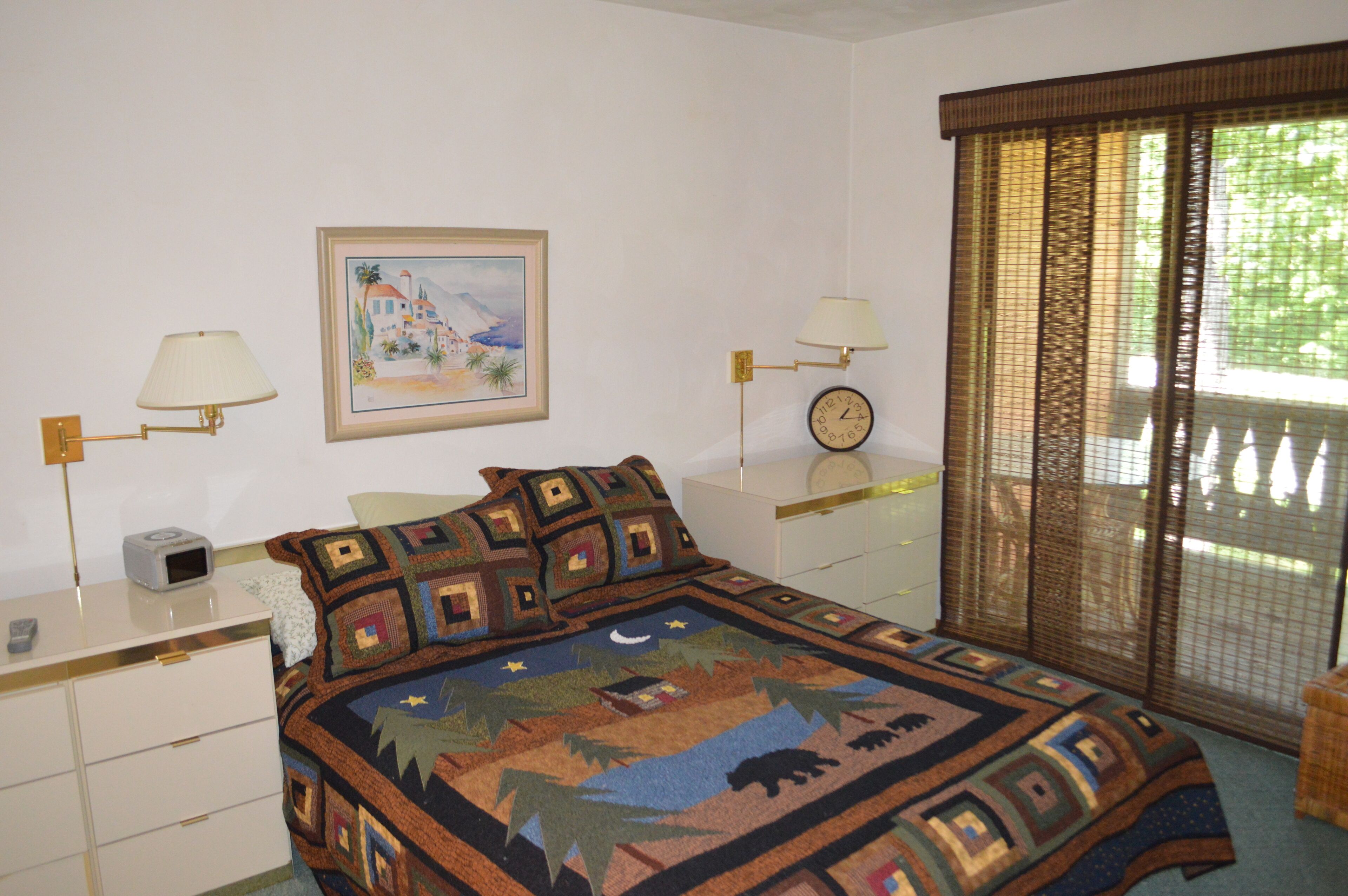 Un dormitorio con cama, dos mesitas de noche, una lámpara, un reloj, un cuadro y una ventana con vistas.