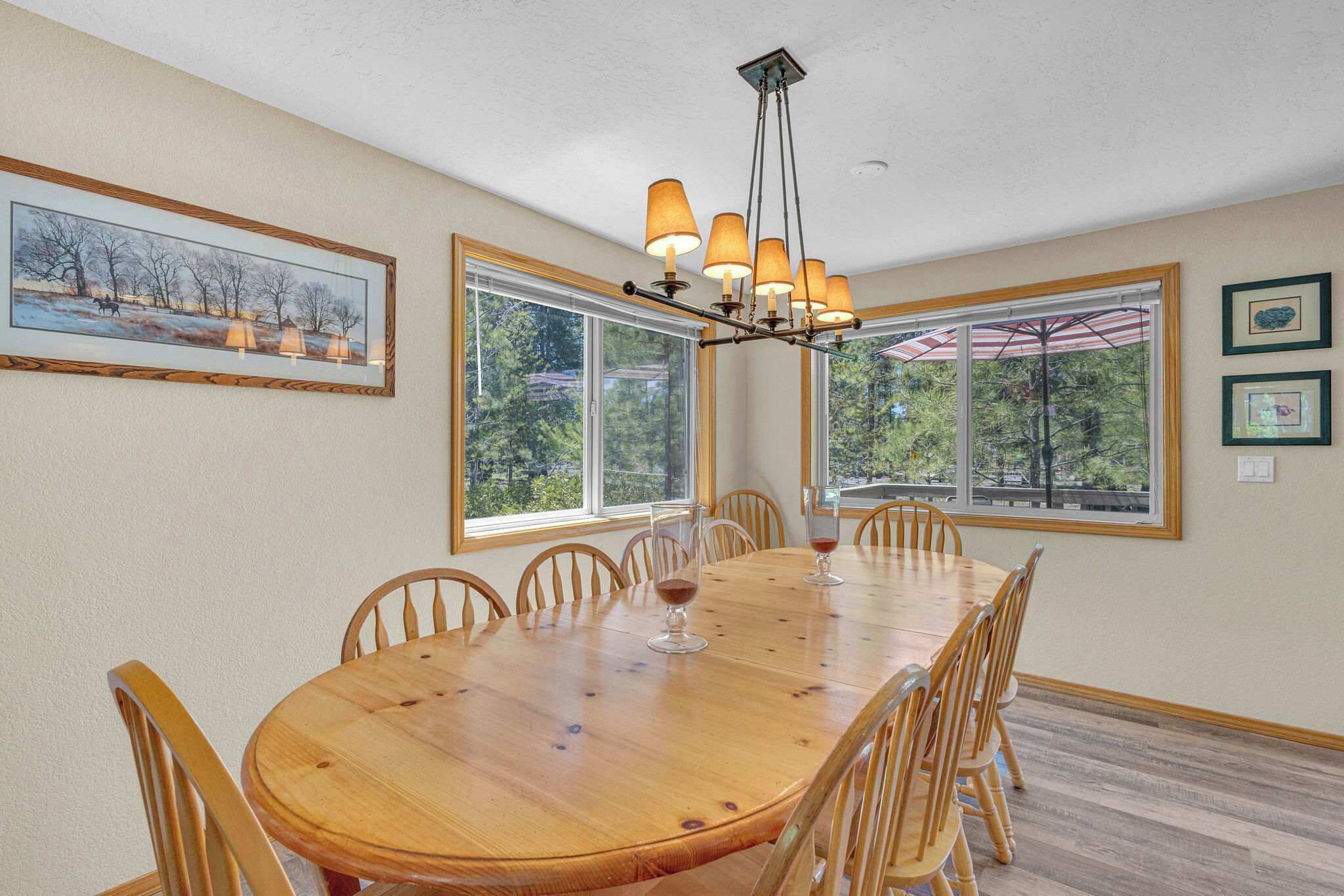 Une salle à manger avec une table en bois, des chaises et un lustre. De grandes fenêtres offrent une vue sur les arbres et un parasol est installé sur la terrasse.