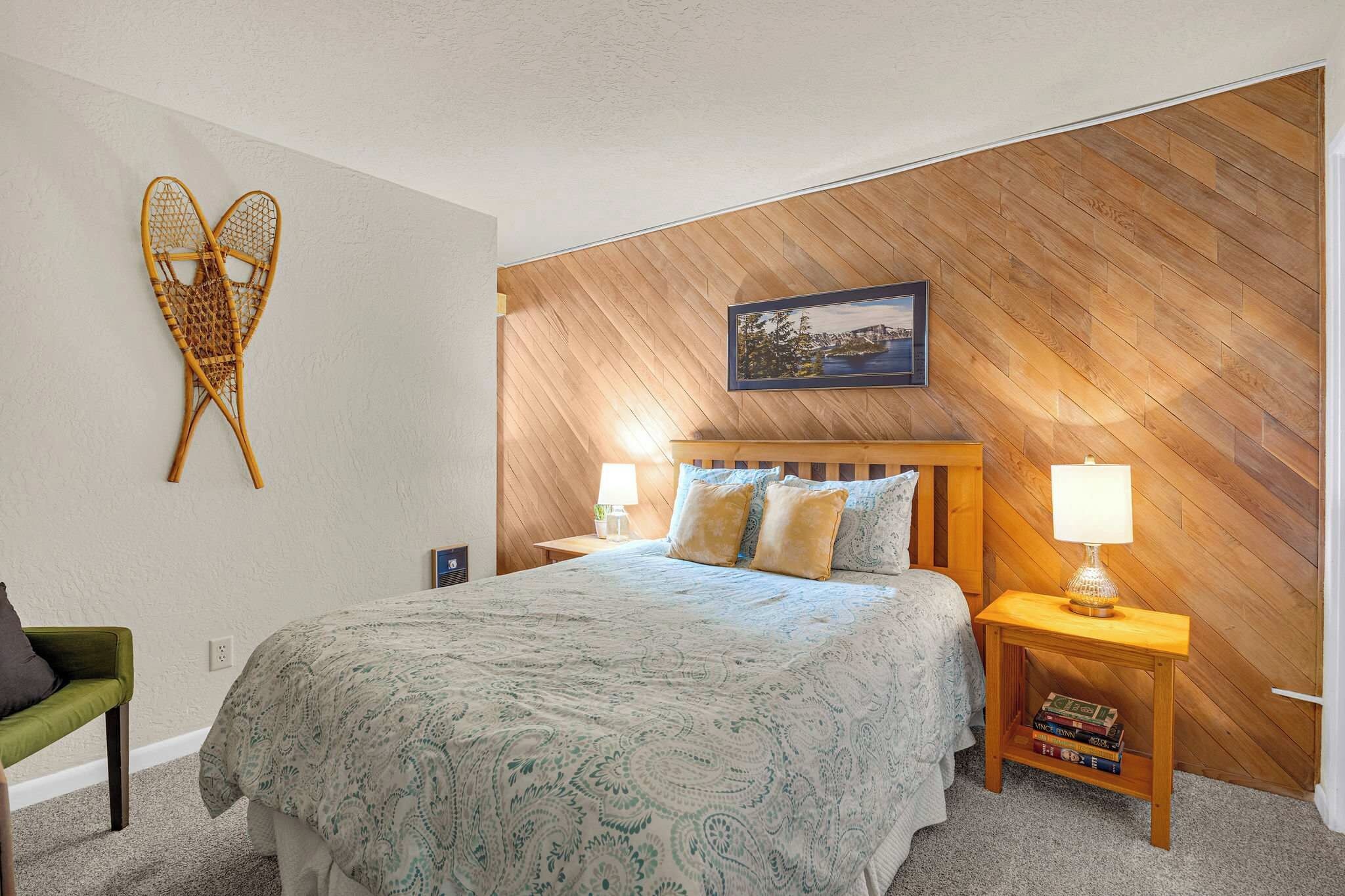 A bedroom with a bed, two bedside tables, a green chair, a wall-mounted picture, and a wooden accent wall.
