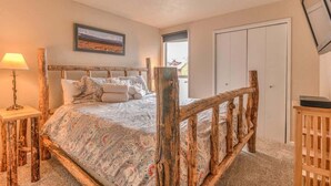 A bedroom with a rustic wooden bed frame, a bedside table with a lamp, a framed picture on the wall, and a door. - Inside Sunriver Village near local shops, eateries (Sunriver)