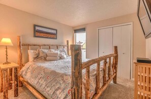 A bedroom with a rustic wooden bed frame, a bedside table with a lamp, a framed picture on the wall, and a door. - Inside Sunriver Village near local shops, eateries (Sunriver)