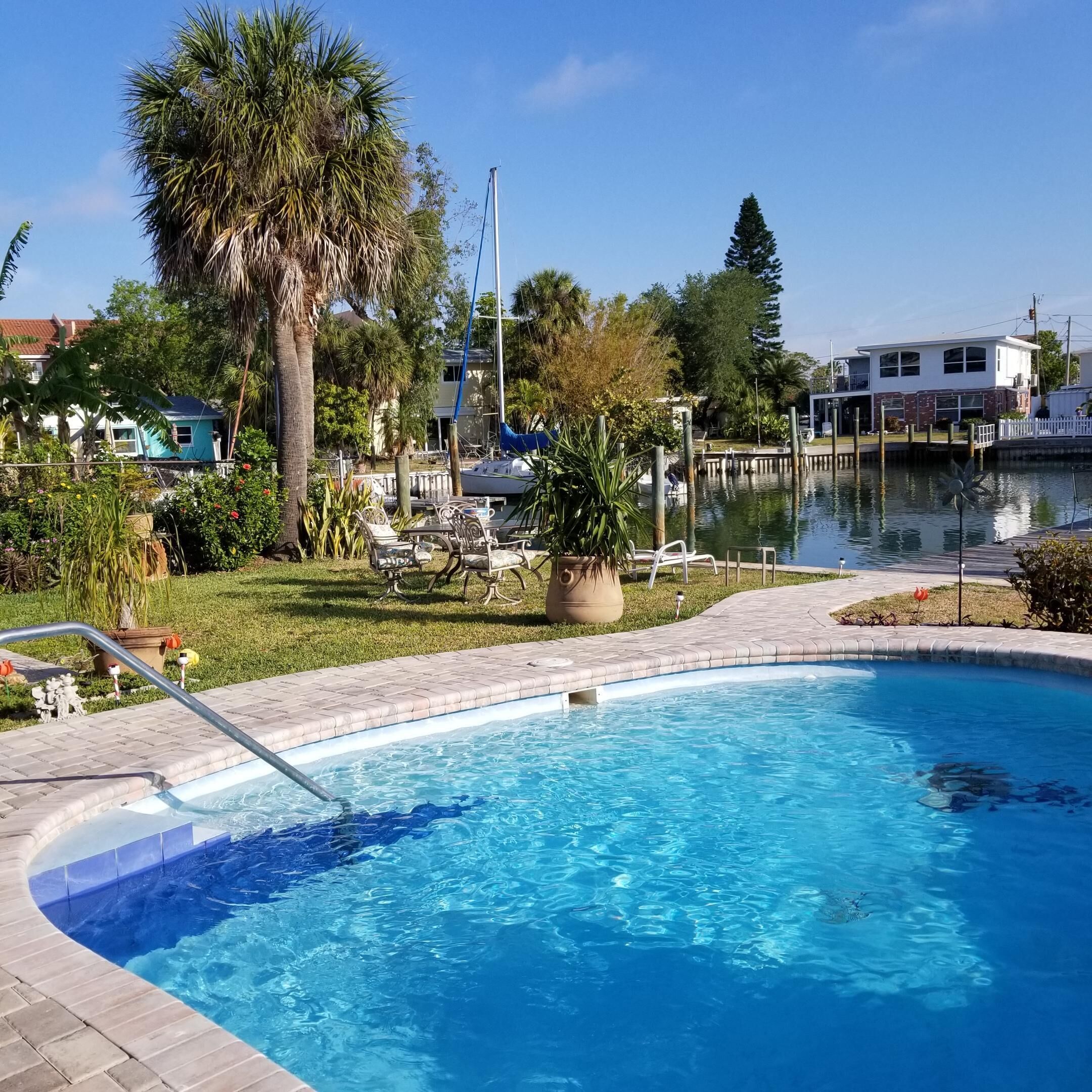 A swimming pool with a clear view of the water, surrounded by a grassy area with chairs and a palm tree.